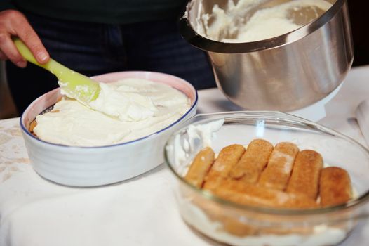 Close-up. Chef's hand putting mixed mascarpone and whipped cream cheese cream on the cake while making tiramisu - traditional Italian dessert at home. Step-by-step recipe