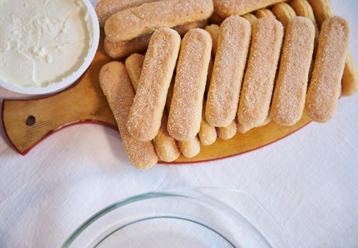 Flat lay composition of savoiardi biscuit cookies on a wooden board and a bowl with mascarpone cheese for The Italian dessert Tiramisu. Copy ad space for text. Step-by-step recipe