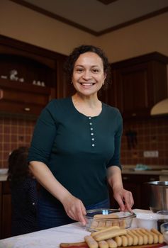 Multiethnic pastry chef woman smiles looking at camera while standing at kitchen table and preparing homemade traditional Italian dessert Tiramisu. Cooking at home, home food and culinary concept.