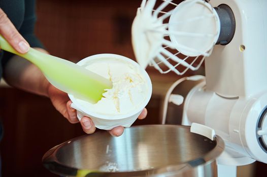 Close-up of a pastry chef's hands using a silicone spatula to put some mascarpone cheese into a steel bowl against a blurred food processor whisk. Preparing the traditional Italian dessert Tiramisu
