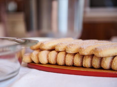 Close-up of ladyfinger savoiardi biscuit cookies on a wooden cutting board on white tablecloth background with blurred steel bowl and glass plate. Close up