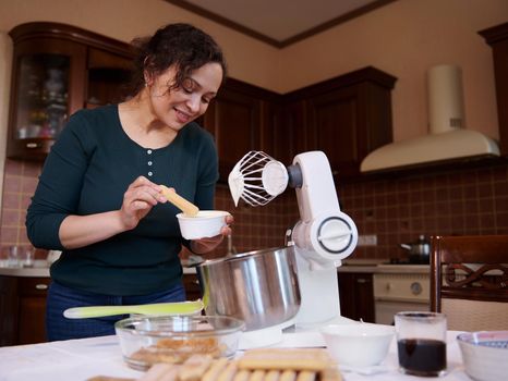 Pretty woman, pastry chef, housewife preparing traditional Italian dessert Tiramisu cake with whipped cream on food processor, mascarpone cheese and ladyfingers biscuit cookies at home kitchen.