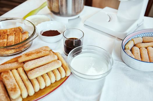 Ingredients for making the traditional Italian sweet tiramisu cake. Savoiardi cookies, strong sweet espresso coffee, cocoa powder, mascarpone cheese and fresh cream on a table with a white tablecloth