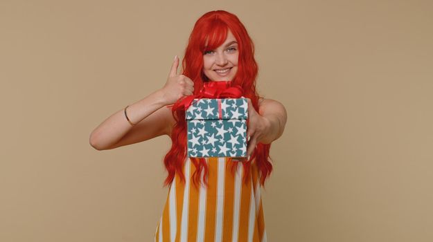 Smiling redhead young woman presenting birthday gift box stretches out hands, offer wrapped present career bonus, celebrating party. Red hair ginger girl isolated alone on beige studio background