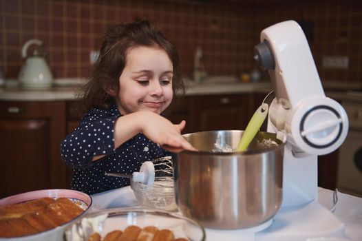 Sweet Caucasian baby girl smiles and makes funny face while licking leftovers of delicious whipped cheese cream from steel bowl, standing at a kitchen table with Tiramisu cake on blurred foreground