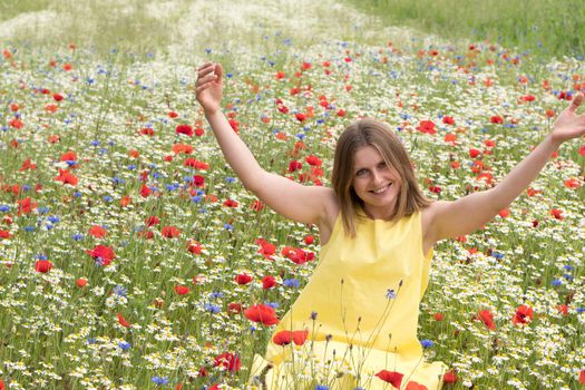 a beautiful young blonde woman in a yellow dress stands among a flowering field of poppies, daisies, cornflowers and laughs. High quality photo