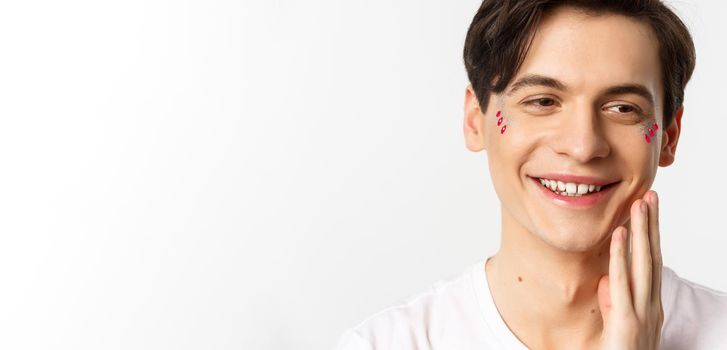 People, lgbtq and beauty concept. Headshot of beautiful gay man with glitter on face, smiling and looking happy, touching cheek after kiss, white background.