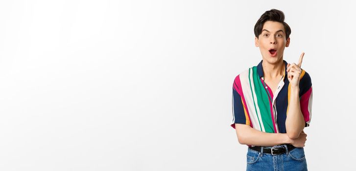 Portrait of young handsome guy having an idea, raising finger and saying suggestion, standing over white background.