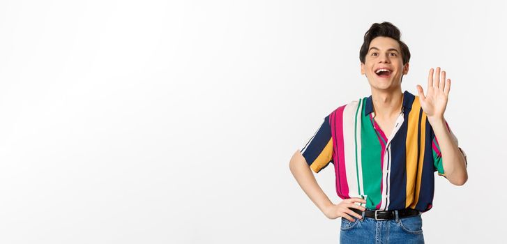 Cheerful gay man saying hello, waving hand and greeting person, standing over white background.