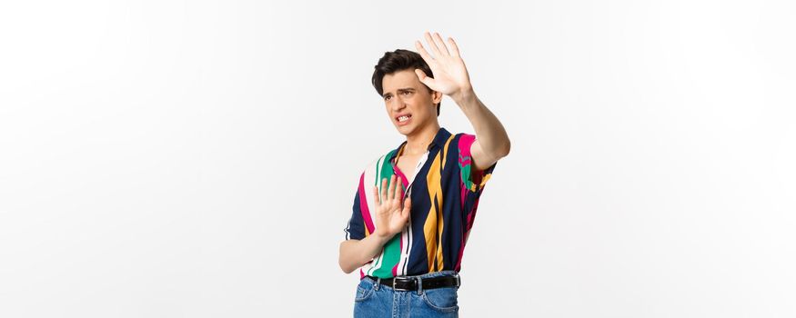Timid and displeased androgynous man asking to stop, raising hands defensive and grimacing, standing over white background.