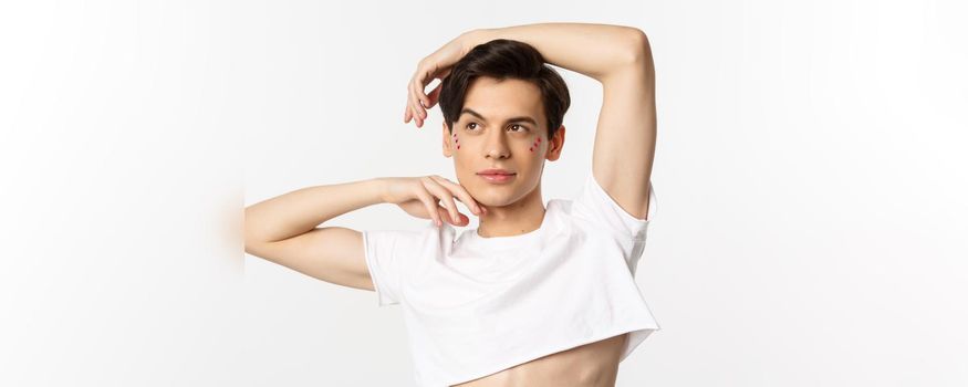 Close-up of beautiful androgynous man in crop top posing for camera, standing against white background.
