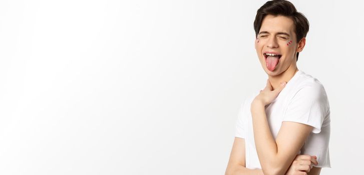 People, lgbtq and beauty concept. Close-up of carefree gay man with glitter on face, showing tongue and making funny face, standing over white background.