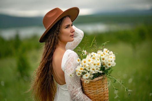 A middle-aged woman in a white dress and brown hat stands on a green field and holds a basket in her hands with a large bouquet of daisies. In the background there are mountains and a lake