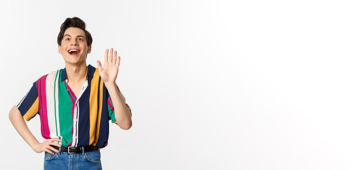 Cheerful gay man saying hello, waving hand and greeting person, standing over white background.