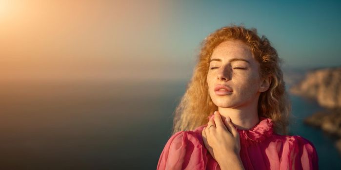 Side view a Young beautiful sensual woman in a red long dress posing on a volcanic rock high above the sea during sunset. Girl on the nature on overcast sky background. Fashion photo