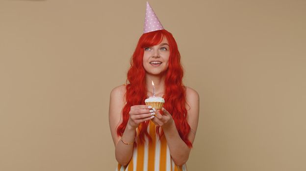 Happy redhead young woman in festive cone cap celebrating birthday anniversary party, blowing candle on small cake cupcake making a wish. Red hair ginger girl isolated alone on beige studio background