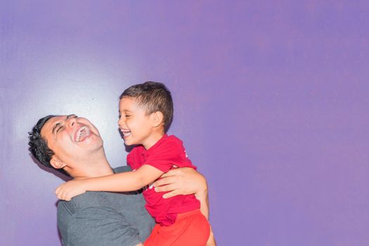 Latin dad smiling and carrying his son in his arms on flat purple background