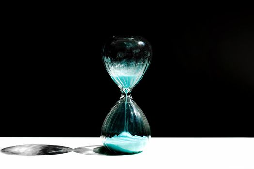 Hourglass, hourglass with blue sand on a black background with a streak of sunlight and a shadow