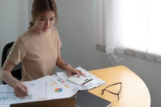 Beautiful Asian businesswoman analyzes charts using laptop calculator at the office..