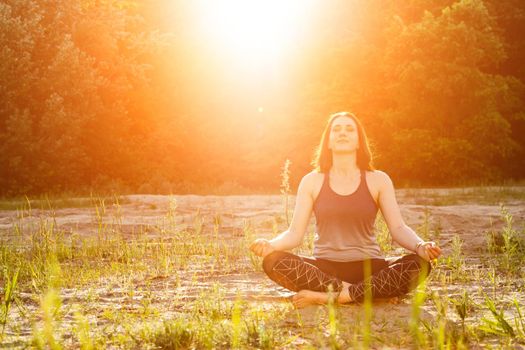 a woman practices yoga and meditates in the lotus position on the sand against the backdrop of trees.