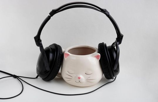 Black headphones are worn on a white coffee mug in the shape of a cat with pink ears, close-up on a white background.