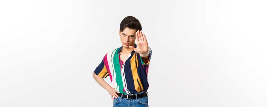 Serious young man extend hand, showing stop gesture, disapprove and prohibit action, standing over white background.
