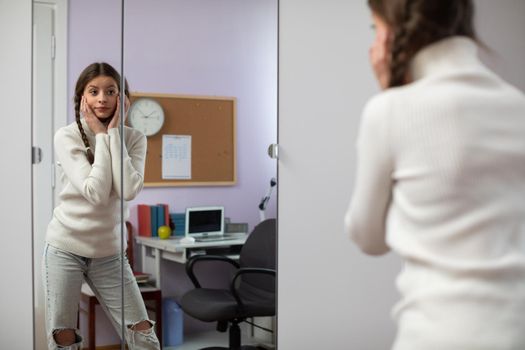 Young brunette girl poses in front of large mirror in room and changes facial expressions. Attractive young girl with two braids.