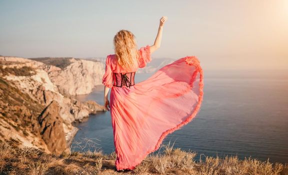 Side view a Young beautiful sensual woman in a red long dress posing on a volcanic rock high above the sea during sunset. Girl on the nature on blue sky background. Fashion photo