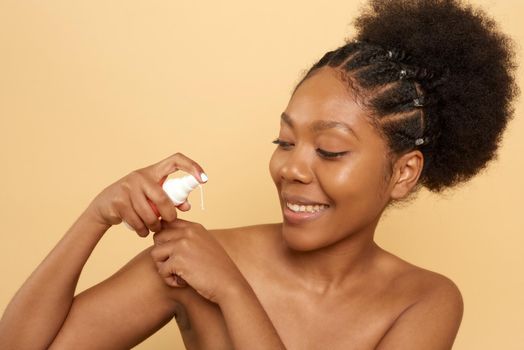 Dark-skinned woman with a wide smile, holds a moisturizer near her face, enjoys the softness of the skin, isolated on a beige background. Facial treatments.