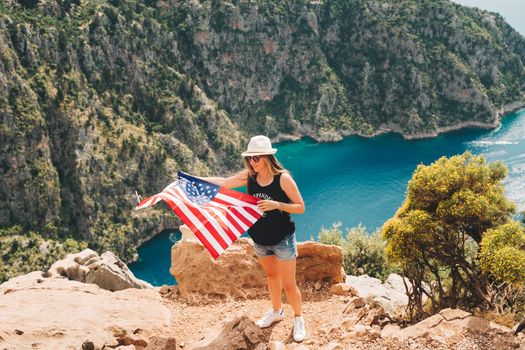 Young woman standing on a rock cliff and waving the US flag while looking at sea beneath. Girl traveller waving the American flag while standing on a mountain top. 4 July Independence day