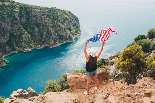 Young woman standing on a rock cliff and waving the US flag while looking at sea beneath. Girl traveller waving the American flag while standing on a mountain top. 4 fourth July Independence Day