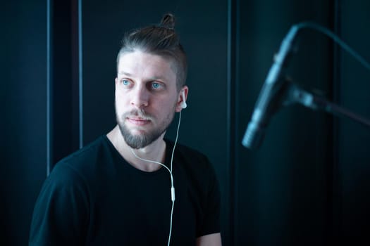 Portrait of a caucasian bearded musician in headphones in a black recording studio with a microphone.