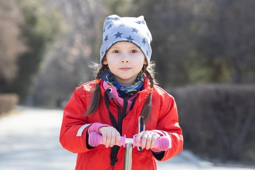 Portrait of a caucasian girl 5 -7 years old on a scooter in a spring park.