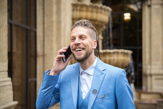 A young man with a hipster hairstyle in a business suit happily speaks on the phone.