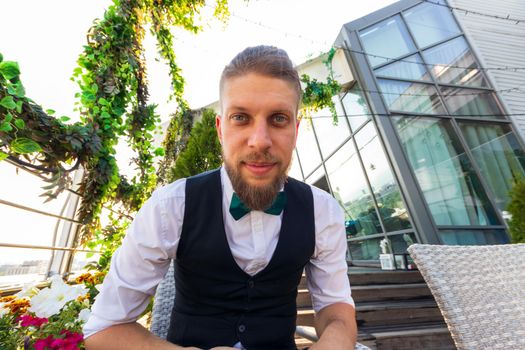Young bearded man in a white shirt, black vest and bow tie looks at the camera while sitting in a penthouse.