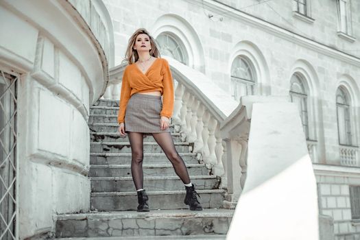 A middle-aged woman looks like a good blonde with curly beautiful hair and makeup on the background of the building. She is wearing a yellow sweater