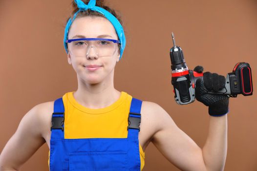 Gender equality. Portrait of a young smiling woman in a uniform working in a workshop, holding an electric drill screwdriver in her hands. Studio shot on brown background.