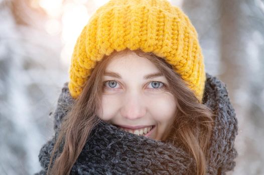 Close-up portrait of a smiling young caucasian woman in a bright woolen hat and scarf, against the backdrop of a snowy forest