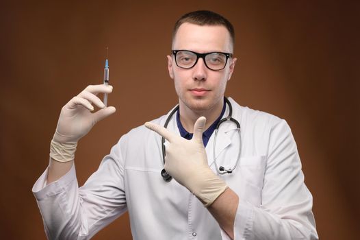 A young Caucasian doctor in a white coat and glasses holds a syringe with a vaccine. Friendly cuddly young man points a finger at a syringe offering to be vaccinated.