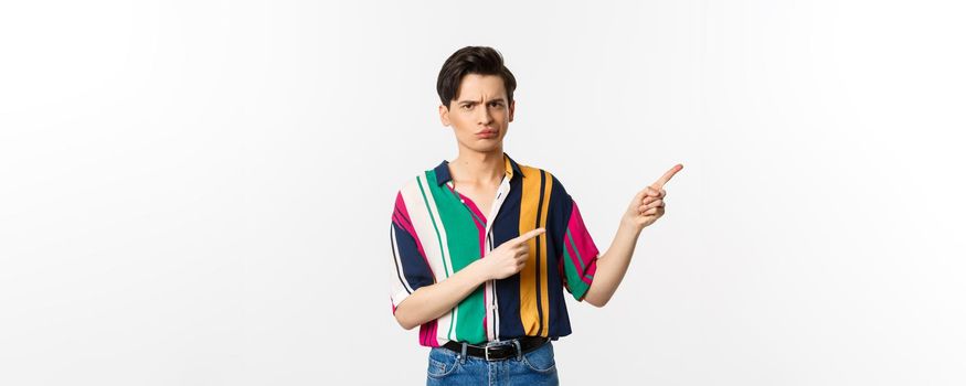 Skeptical and displeased young man having concern, poitning fingers right and looking with disapproval, standing over white background.
