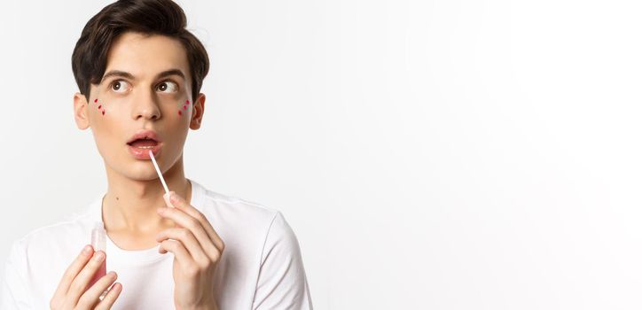 Close-up of beautiful androgynous man with glitter under eyes applying lip gloss, looking dreamy at upper left corner, white background.