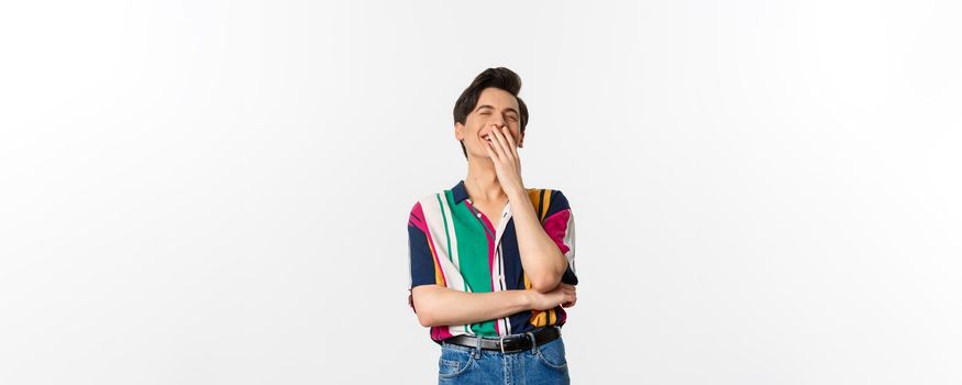 Image of handsome young man laughing over joke, having fun, cover mouth with hands and chuckle, standing over white background.