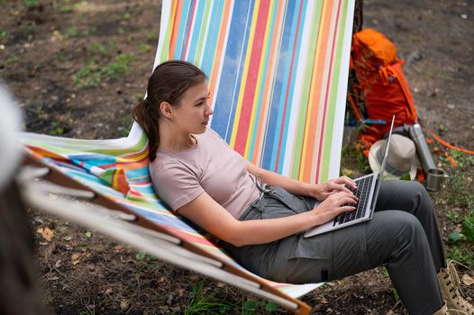 Caucasian woman working on laptop while sitting in a hammock in the forest. Girl uses a wireless computer on a hike