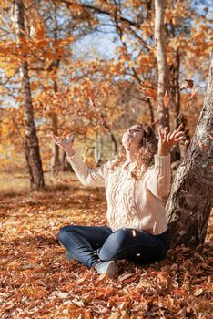 Autumn nature. Happy young woman throwing yellow leaves up in the air sitting in the autumn forest