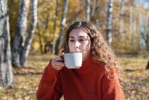 Leisure, free time. Beautiful caucasian woman in red sweater on a picnic outdoors, sitting on a plaid in autumn forest