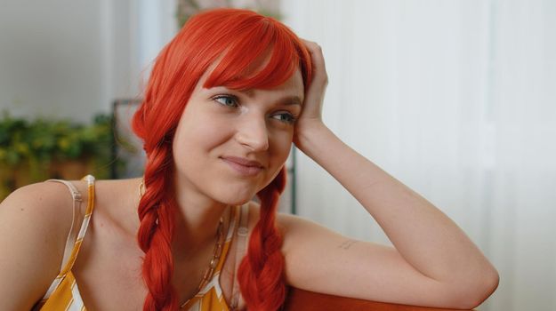 Close up of beautiful happy calm young caucasian redhead girl smiling friendly, glad expression looking away dreaming resting, relaxation feel satisfied concept good news sitting on sofa at home room