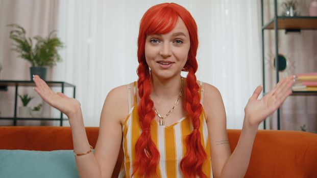 POV of redhead young woman sitting on couch, looking at camera, making video webcam conference call with friends or parents, enjoying pleasant conversation. Girl laughing, waving hello alone in home