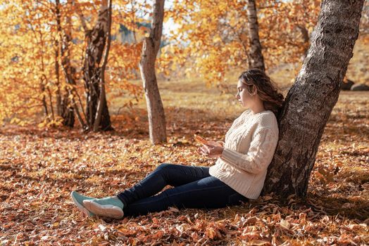 Autumn nature. Young thoughtful woman sitting by the tree in autumn forest