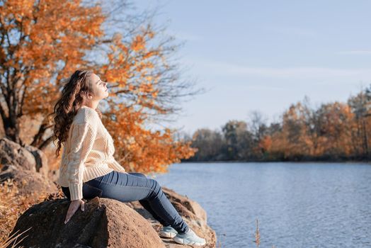 Freedom concept. Thoughtful romantic woman sitting on rocks on the river bank in sunset in autumn day, copy space.