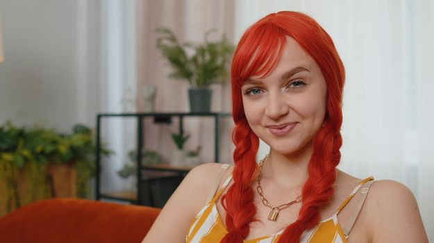 Close-up portrait of happy redhead teenager redhead girl in casual tank top smiling, looking at camera. Young fashion ginger woman indoor isolated at home in living room on couch. Female nature beauty